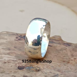 May include: A polished silver ring with a rounded band. The ring reflects the surrounding environment, creating a mirrored effect. The ring is set on a wooden surface. The text "925BangleShop" is visible at the bottom of the image.