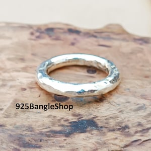 Pode incluir: Um anel de prata com textura martelada. O anel é circular e tem uma superfície ligeiramente irregular. O anel é exibido em uma superfície de madeira. O texto "925BangleShop" é visível.