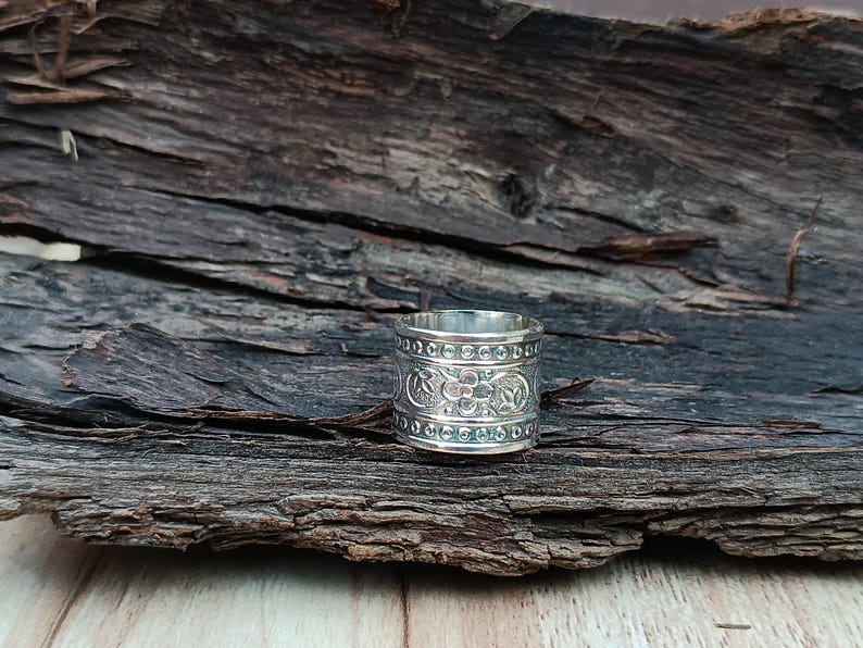 K&ouml;nnte beinhalten: Ein silberner Ring mit einem komplizierten floralen Design. Der Ring ist breit und hat eine strukturierte Oberfl&auml;che. Er liegt auf einem St&uuml;ck verwittertem Holz.