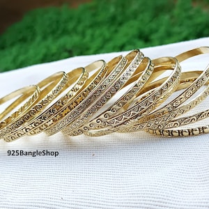 May include: A selection of gold-coloured bangles with detailed engraved patterns. The bangles are displayed on a white surface, with a blurred green backdrop. The bangles feature diverse designs, including floral and geometric patterns.