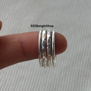 Puede incluir: Un conjunto de anillos apilables de plata. Los anillos están sujetos por un dedo. Los anillos tienen un acabado pulido con un diseño texturizado en el anillo central. El texto "925BangleShop" es visible.