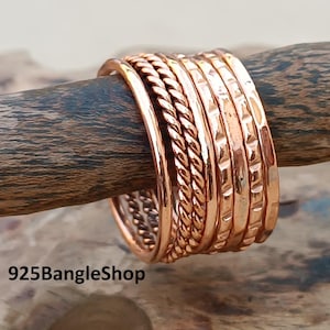 May include: A stack of copper-colored rings with various textures, including smooth, braided, and hammered designs. The rings are displayed on a wooden branch, showcasing their warm metallic tones. The rings are a jewelry item.