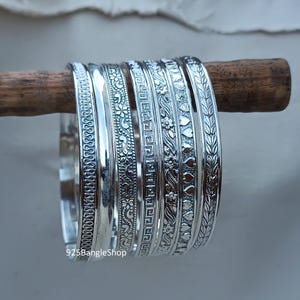 Op de afbeelding: Set van zes zilveren armbanden met verschillende gegraveerde ontwerpen, waaronder bloemen, geometrische en bladpatronen. De armbanden zijn gestapeld op een houten oppervlak. 925BangleShop is gedrukt onderaan de afbeelding.