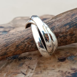 Könnte beinhalten: Ein silberner Ring mit drei ineinandergreifenden Bändern. Der Ring hat eine polierte, reflektierende Oberfläche und ist vor einem natürlichen Holzuntergrund platziert. Das Design des Rings ist schlicht, aber elegant.