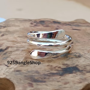 May include: A silver-colored ring in a spiral design. The ring wraps around the finger with a smooth, polished finish. The text "925BangleShop" is visible at the bottom of the image. The ring is displayed on a wooden surface.