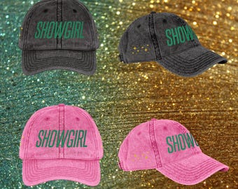 May include: Four baseball caps are shown. Two are dark grey, and two are pink. Each cap features the word "SHOWGIRL" or "SHOW" in green lettering. Some caps have gold star accents. The caps are made of a distressed fabric.