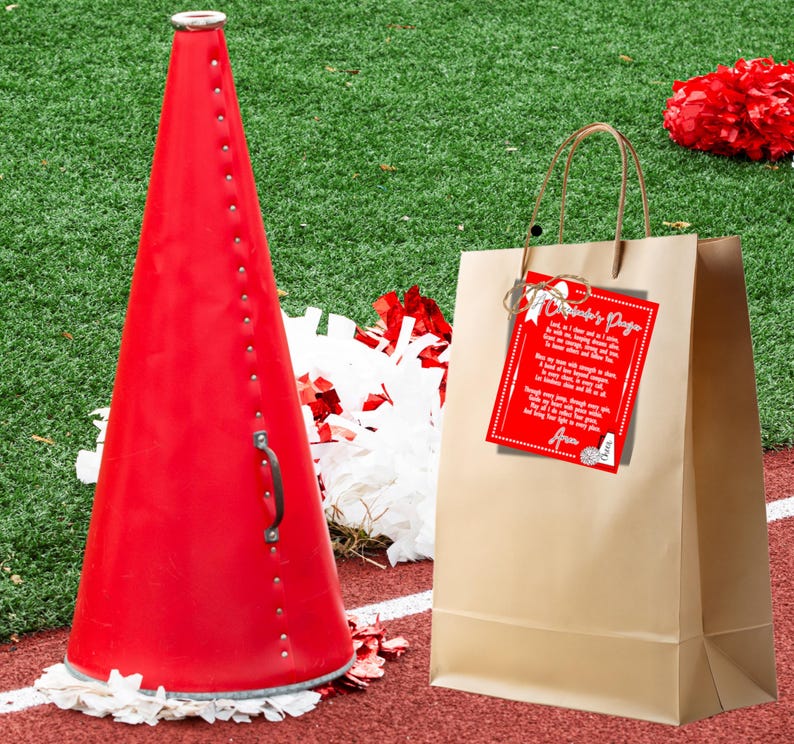 May include: A red megaphone with a white handle, a brown paper bag with a red and white gift tag that says "Cheerleader's Prayer", and white and red pom-poms on a green artificial grass surface.