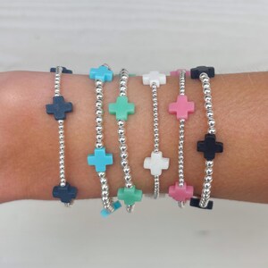 May include: Six beaded bracelets with cross-shaped charms in various colors: navy, turquoise, green, white, pink, and black. The bracelets feature silver-colored beads and are displayed on a wrist.