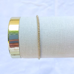 May include: A gold beaded bracelet on a white fabric display stand.