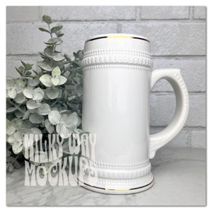 May include: White ceramic beer stein with a gold rim and a textured design. The stein has a handle and is sitting on a marble surface.