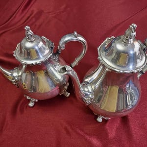 Op de afbeelding: Twee zilveren theepotten met sierlijke ontwerpen en handvatten. De theepotten hebben een gepolijste afwerking en staan op een rood oppervlak.