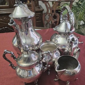 May include: A silver tea set with decorative handles and lids. The set includes a teapot, coffee pot, sugar bowl, creamer, and a small serving bowl. The pieces are arranged on a red tablecloth.