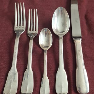 May include: A set of five silver-colored antique flatware pieces arranged on a burgundy surface. The set includes two forks, two spoons, and a knife. The flatware has a classic design with a shiny finish. The letters A, B, C, D, and E are at the bottom.