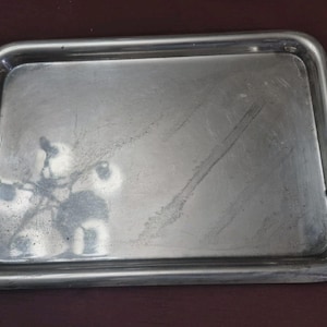 Puede incluir: Una bandeja rectangular de acero inoxidable con esquinas redondeadas. La bandeja tiene una superficie ligeramente reflectante con algo de desgaste visible. Los bordes están elevados, formando un borde alrededor del perímetro. La bandeja está diseñada para transportar y servir alimentos o bebidas.