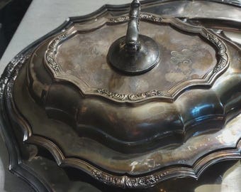Könnte beinhalten: Eine silberfarbene Servierschale mit Deckel und Griff. Die Schale hat einen dekorativen, gewellten Rand und aufwendige Details. Der Deckel hat einen zentralen Griff und ein Monogramm. Die Schale ist wahrscheinlich aus Metall.