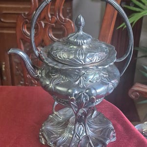 May include: An antique silver teapot and stand. The teapot has a floral pattern on the lid and body, with a curved handle. The stand has a decorative, leaf-like base. The set is displayed on a red surface.