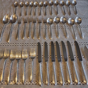 May include: A set of vintage silverware, including spoons, forks, and knives, arranged on a patterned tablecloth. The silverware has a shiny, silver finish. The set includes various sizes of spoons and forks, along with several knives.