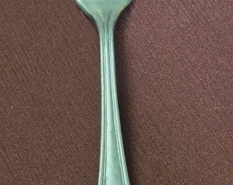 May include: A silver-colored fork with four tines, set against a brown background. The fork's handle tapers to a rounded end. The utensil is likely for dining.