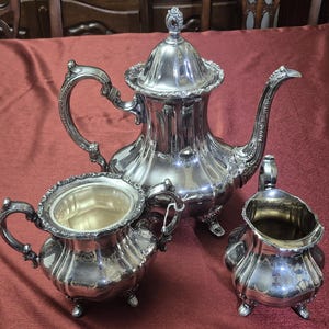 May include: A silver tea set with a teapot, creamer, and sugar bowl. The set is ornate with intricate details and a polished finish.