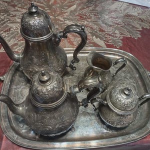 May include: A silver tea set with ornate floral detailing, including a teapot, a creamer, a sugar bowl, and a second teapot, all resting on a matching tray. The set has a vintage aesthetic.