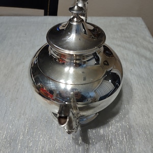 May include: A silver teapot with a shiny, polished finish. The teapot has a round body and a lid with a knob on top. The spout is curved and has a decorative design.