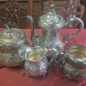 May include: A silver tea set with ornate floral detailing. The set includes a teapot, sugar bowl, creamer, and a covered serving dish. The pieces are arranged on a red tablecloth, with a wooden chair in the background.