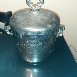 May include: A silver ice bucket with a decorative lid and handles. The lid has a ornate design with a raised circle in the center.