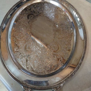 May include: A silver oval serving tray with two handles. The tray has an ornate design with a raised rim.