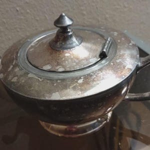 May include: A vintage silver teapot with a rounded body, spout, and handle. The lid has a small finial and a hinged opening. The teapot shows signs of age and wear, with some discoloration on the surface. A classic piece for tea lovers.