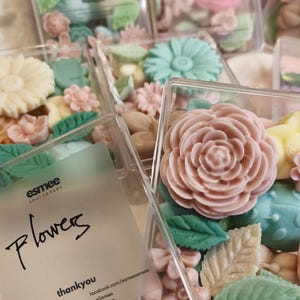 Puede incluir: Surtido de pequeños jabones con forma de flores de varios colores en recipientes de plástico transparente. Los jabones están dispuestos en una variedad de colores, incluyendo rosa, blanco, verde y azul. Los recipientes están etiquetados con el texto "esmee soul senses Flowers".