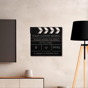 May include: A black movie clapperboard with white chevron accents. The clapperboard has text that reads "YOUR CUSTOM WORDS" and "A UNIQUE 2U PRODUCTION". The date "8 17 2025" is also displayed.