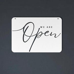 May include: A rectangular white sign with rounded corners, featuring the words "WE ARE Open" in elegant black script. The sign is set against a dark gray background, creating a high-contrast visual.
