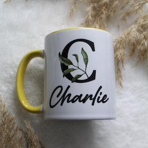 Op de afbeelding: Witte keramische mok met een gele rand en handvat. De mok heeft een zwarte letter "C" met groene bladeren en de naam "Charlie" in zwarte cursieve letters.