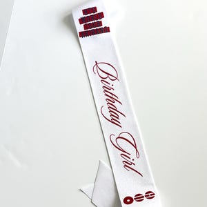 May include: A white satin sash featuring the red text "Birthday Girl" in a cursive script. The sash includes a red graphic element at the bottom consisting of three circular icons and a red rectangular block with indistinct text at the top.