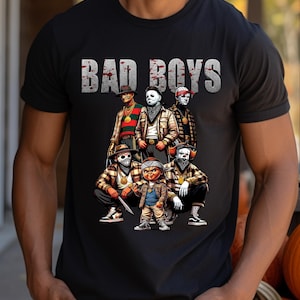 Man wearing a black Halloween t-shirt featuring the Bad Boys of Horror PNG design with Freddy Krueger, Michael Myers, Pinhead, Jason Voorhees, Pennywise, and Chucky. The characters are dressed in brown plaid streetwear with bandanas, sneakers, and gold chains, standing under bold gray blood-splattered text that reads ‘Bad Boys.’ Horror PNG artwork displayed on apparel.