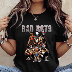 Woman wearing a black Halloween t-shirt featuring the Bad Boys of Horror PNG design with Freddy Krueger, Michael Myers, Pinhead, Jason Voorhees, Pennywise, and Chucky. The characters are dressed in brown plaid streetwear with bandanas, sneakers, and gold chains, standing under bold gray blood-splattered text that reads ‘Bad Boys.’ Horror PNG artwork displayed on apparel.