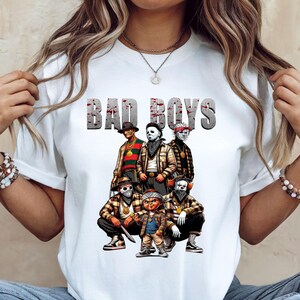 Woman wearing a white Halloween t-shirt featuring the Bad Boys of Horror PNG design with Freddy Krueger, Michael Myers, Pinhead, Jason Voorhees, Pennywise, and Chucky. The characters are dressed in brown plaid streetwear with bandanas, sneakers, and gold chains, standing under bold gray blood-splattered text that reads ‘Bad Boys.’ Horror PNG artwork displayed on apparel.