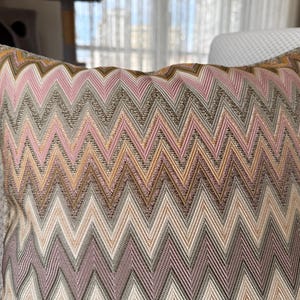 May include: Decorative pillow with a zig-zag pattern in shades of pink, brown, and beige. The pillow has a textured appearance and is placed on a light-colored surface. The pillow's design adds a touch of elegance to home decor.