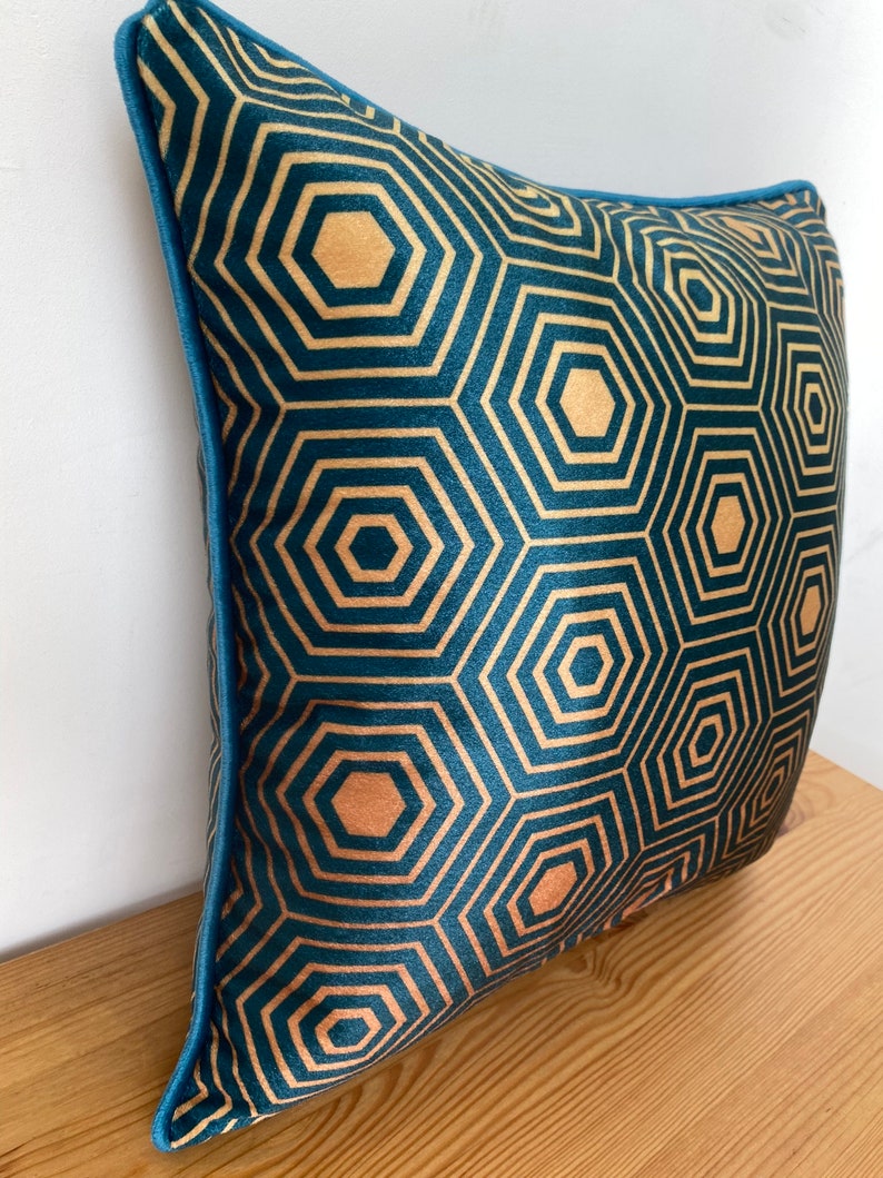 Puede incluir: Una almohada de terciopelo azul turquesa con un patr&oacute;n geom&eacute;trico dorado de hex&aacute;gonos. La almohada tiene un ribete azul turquesa.