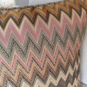 May include: Close-up of a decorative pillow with a woven chevron pattern. The design features a repeating zigzag pattern in shades of pink, gold, gray, and brown. The pillow has a textured appearance and a soft, inviting look.