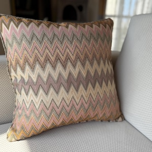Könnte beinhalten: Ein dekoratives quadratisches Kissen mit einem Zickzackmuster in Rosa-, Beige- und Brauntönen. Das Kissen liegt auf einem weißen Sofa. Die Kanten sind mit einem dunkleren Stoff besetzt.