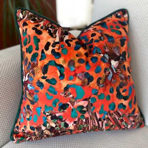 May include: Square decorative pillow with a vibrant orange, teal, and black animal print pattern. The pillow has a dark green trim and is placed on a light gray textured surface. The pillow's design features abstract shapes and colors.