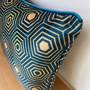 Puede incluir: Una almohada de terciopelo azul verdoso con un patr&oacute;n geom&eacute;trico dorado. La almohada tiene un ribete azul verdoso alrededor de los bordes.