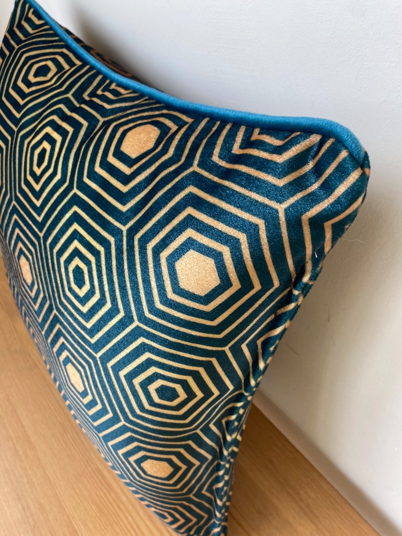 Puede incluir: Una almohada de terciopelo azul verdoso con un patr&oacute;n geom&eacute;trico dorado. La almohada tiene un ribete azul verdoso alrededor de los bordes.