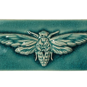 Op de afbeelding: Een turquoise keramische tegel met een reliëf van een cicade-insect. Het insect heeft gedetailleerde vleugels en een lichaam, met een lichtere, gebroken witte kleur. De tegel heeft een gekraakte glazuurafwerking.