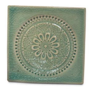 Könnte beinhalten: Eine quadratische Keramikfliese mit hellgrüner Craquelé-Glasur. In der Mitte befindet sich ein erhabenes, kreisförmiges Mandala-Muster mit einem zentralen Blumenmotiv, umgeben von geometrischen Details und einem gepunkteten Rand.
