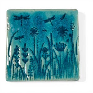 Op de afbeelding: Vierkante keramische tegel met een turquoise achtergrond en een gedetailleerd ontwerp van libellen en wilde bloemen. Het kunstwerk heeft een craquelé-effect, waarbij de libellen en planten in een donkerdere tint turquoise zijn weergegeven.