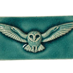 Könnte beinhalten: Eine rechteckige Keramikfliese mit einem erhabenen, dreidimensionalen Relief einer fliegenden Eule mit ausgebreiteten Flügeln. Das Stück hat eine glänzende, blaugrüne Glasur mit einem sichtbaren Craquelé-Finish auf der gesamten Oberfläche.