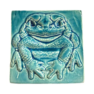 Könnte beinhalten: Quadratische, türkisfarbene Keramikfliese mit einem erhabenen Relief einer Froschdarstellung. Der Frosch hat große Augen und einen breiten Mund. Die Fliese hat eine glänzende Oberfläche. Der Frosch hat eine strukturierte Haut.