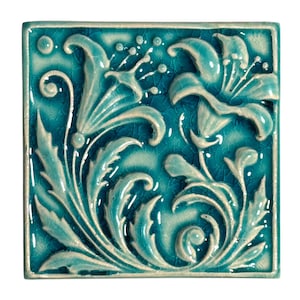Könnte beinhalten: Quadratische türkisfarbene Keramikfliese mit erhabenem Blumendesign. Das Design zeigt stilisierte Lilien und Blätter in einer helleren, cremefarbenen Farbe mit einem Craquelé-Effekt. Die Fliese hat eine glänzende Oberfläche und einen erhabenen Rand.